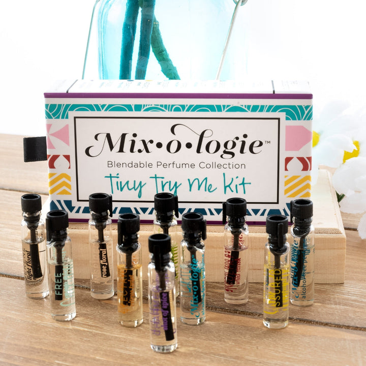 Mixologie Tiny Try Me Kit open slide box propped upright on wood surface with 10 mini perfume vials including Free, Inspired, Sultry, and Assured displayed in front, with teal reed diffuser and white and yellow flowers in background