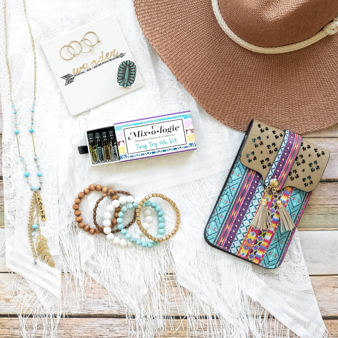 Mixologie Tiny Try Me Kit open slide box flat lay on white lace scarf with gold "Faith" charm necklace, stacked beaded bracelets, boho crossbody bag, straw hat, and gold ring jewelry on white wood background