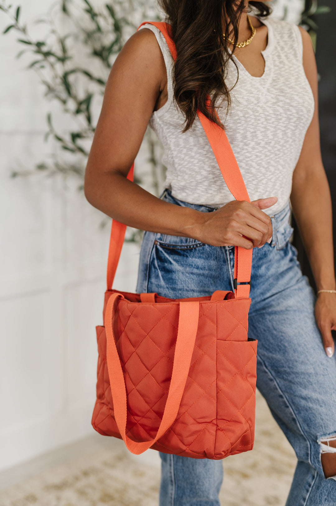 Close-up of a woman carrying the rust orange quilted tote crossbody with a long nylon strap, paired with a white tank and distressed jeans.