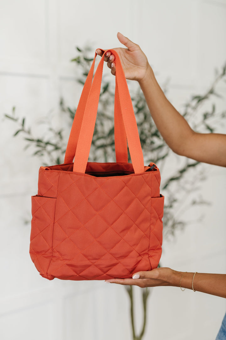 Hands holding up a rust orange quilted diamond-pattern tote bag with flat nylon handles and side pockets, in front of a potted olive tree.
