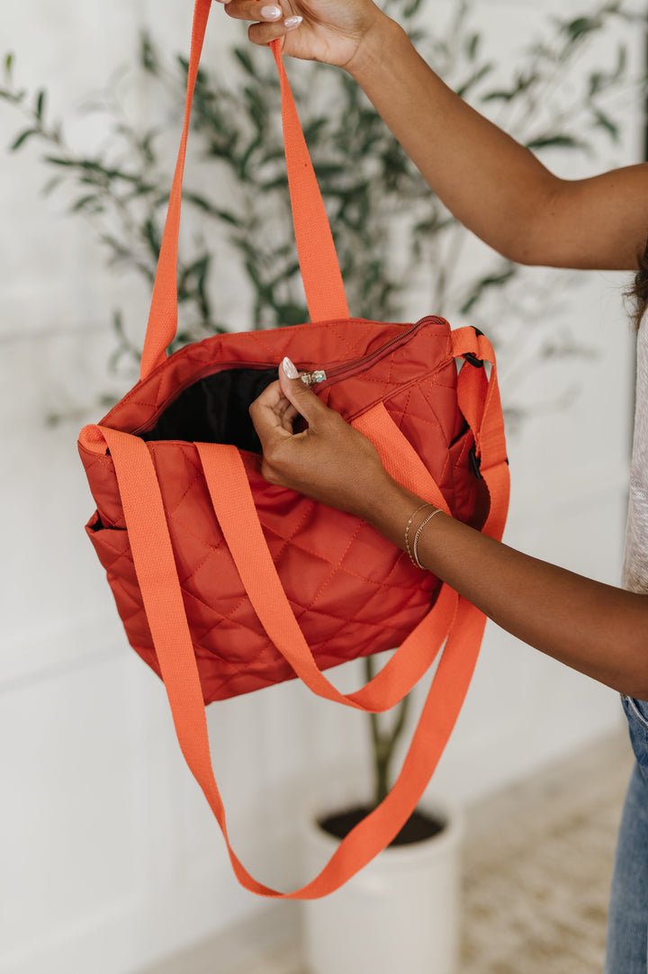 Hands opening the rust orange quilted tote to reveal a dark interior with a zip closure and long crossbody strap.