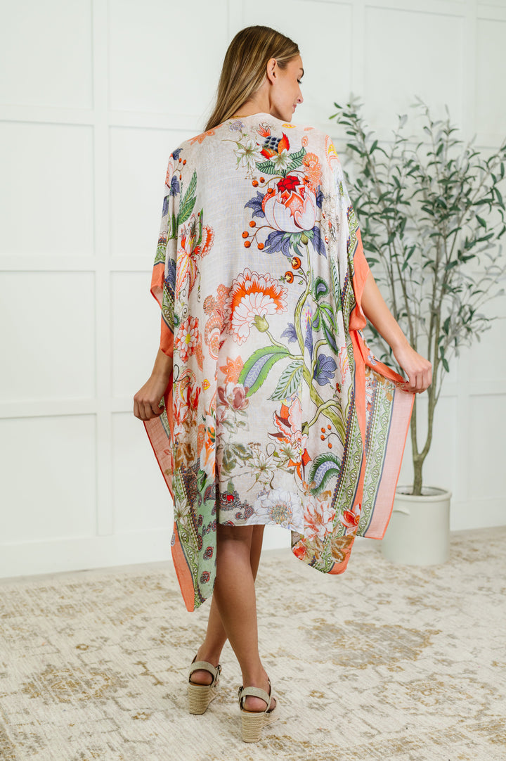 Back view of a woman wearing a floral and paisley print kimono with coral trim, paired with gold sandals, posed in front of a leafy plant