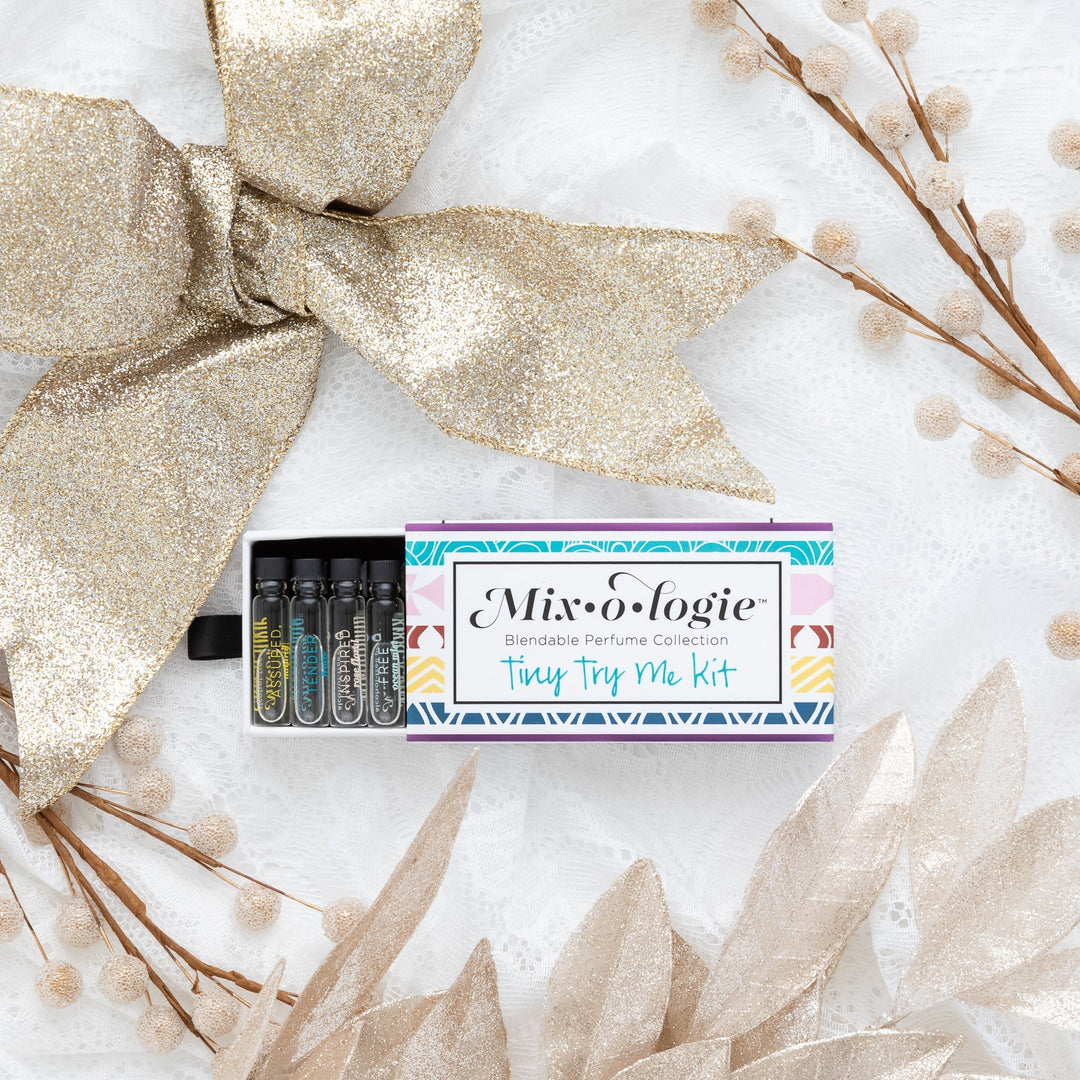 Mixologie Tiny Try Me Kit open slide box showing Assured, Tenacious, Inspired, and Free mini perfume vials, styled flat lay with gold glitter bow, cream poinsettia leaves, and pom pom branches on white background