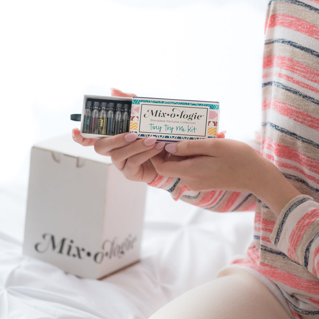 Mixologie Tiny Try Me Kit open slide box held in hands by model in striped top, showing row of mini perfume vials, with white Mixologie gift bag in background