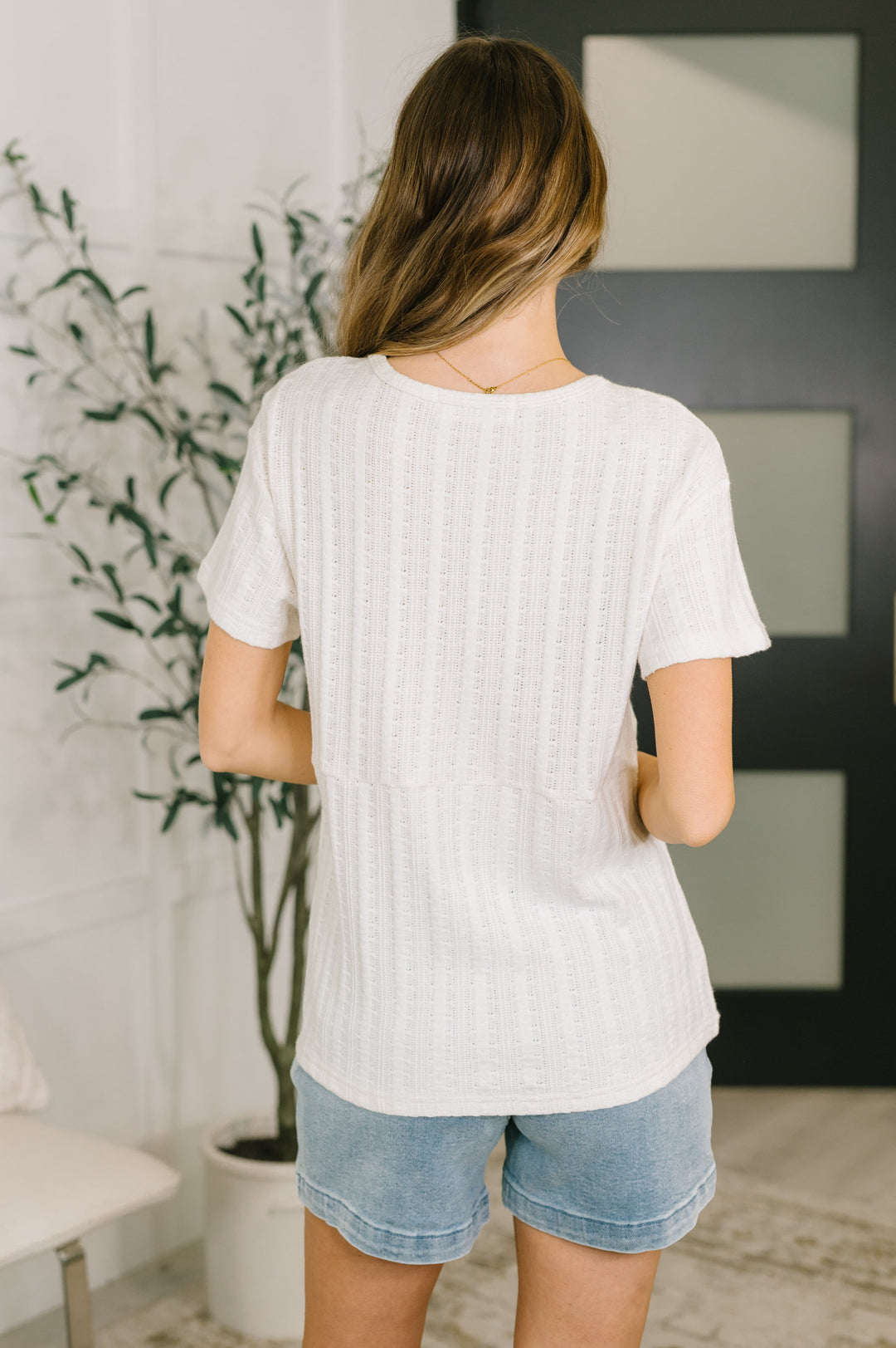 Back view of a woman wearing a cream short-sleeve henley top with denim shorts