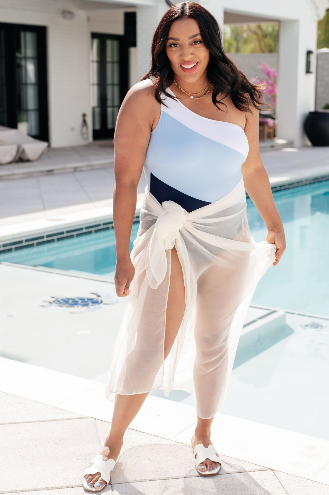 Plus size model wearing a white, light blue, and navy colorblock one-shoulder one-piece swimsuit with a sheer white sarong wrap, styled with white slide sandals poolside.