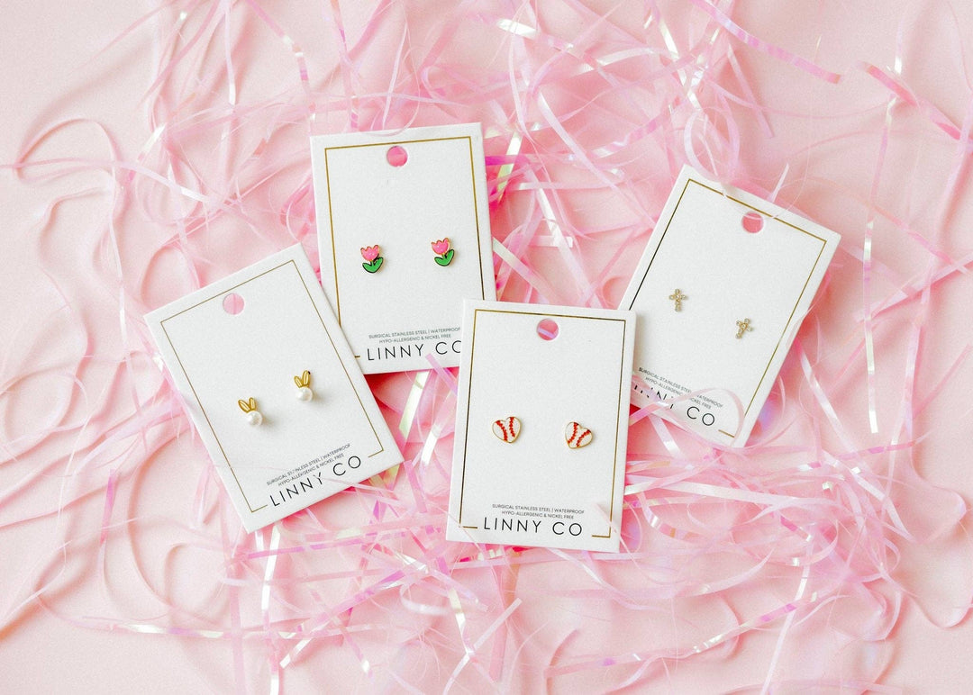 Four Linny Co stud earring cards flat lay on pink ribbon confetti background — gold bunny ear pearl studs, pink tulip enamel studs, red and white baseball enamel studs, and gold cross studs.