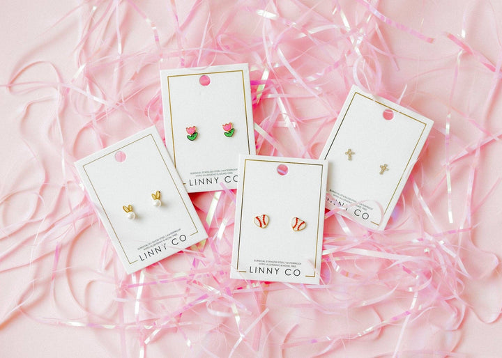 Four Linny Co stud earring cards flat lay on pink ribbon confetti background — gold bunny ear pearl studs, pink tulip enamel studs, red and white baseball enamel studs, and gold cross studs.