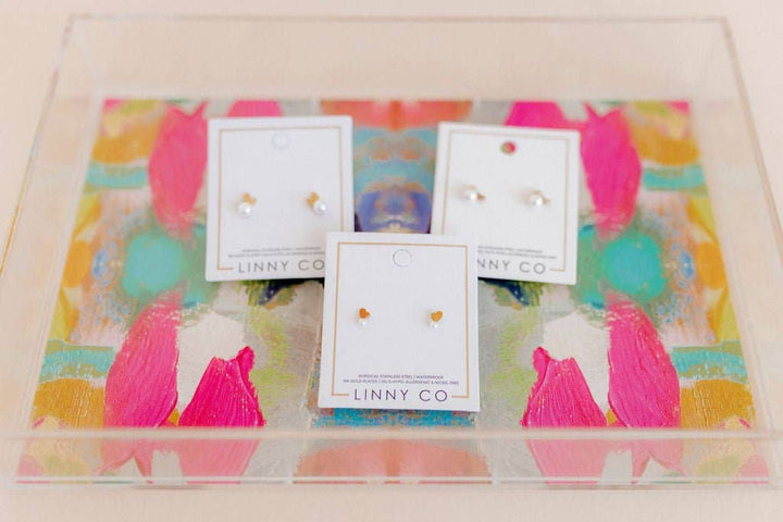 Three Linny Co gold bunny ear and pearl stud earring cards displayed on a colorful abstract painted surface.