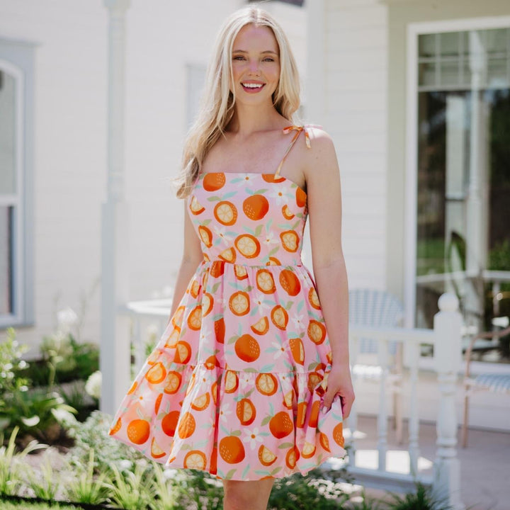 Smiling woman outdoors in a pink orange print tiered mini dress with tie shoulder straps, holding a pink clutch in front of a white house with greenery.