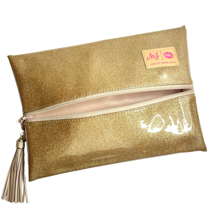 Makeup Junkie Bags gold glitter foldover zip makeup bag partially open showing blush pink interior lining with gold tassel zipper pull and branded patch, front view on white background
