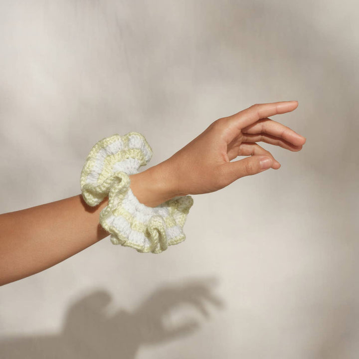 Hand with the butter yellow and white crochet scrunchie worn on the wrist, fingers extended against a neutral background.