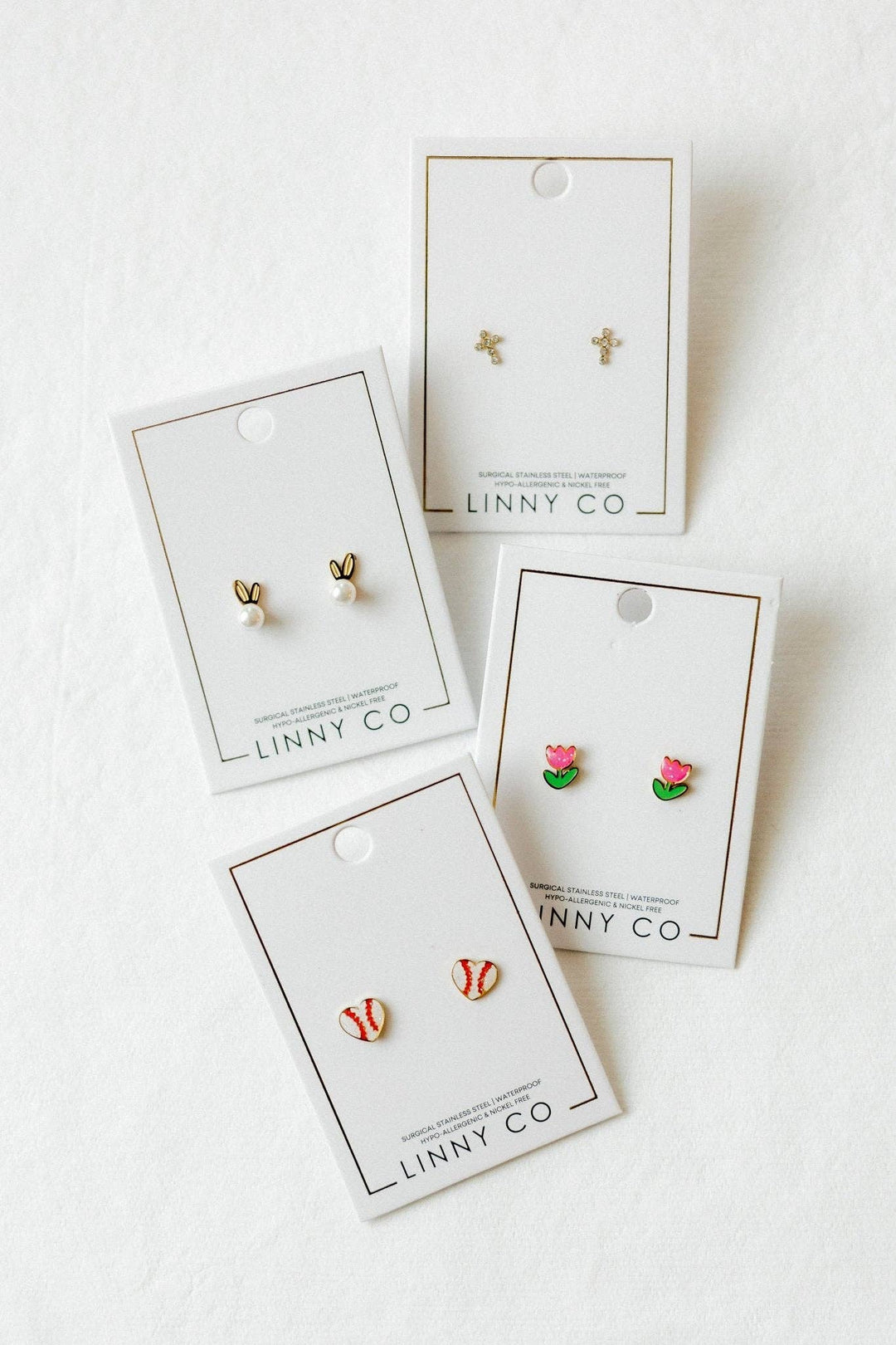 Four Linny Co stud earring cards flat lay on white background — gold bunny ear pearl studs, gold cross studs, pink tulip enamel studs, and red and white baseball enamel studs.