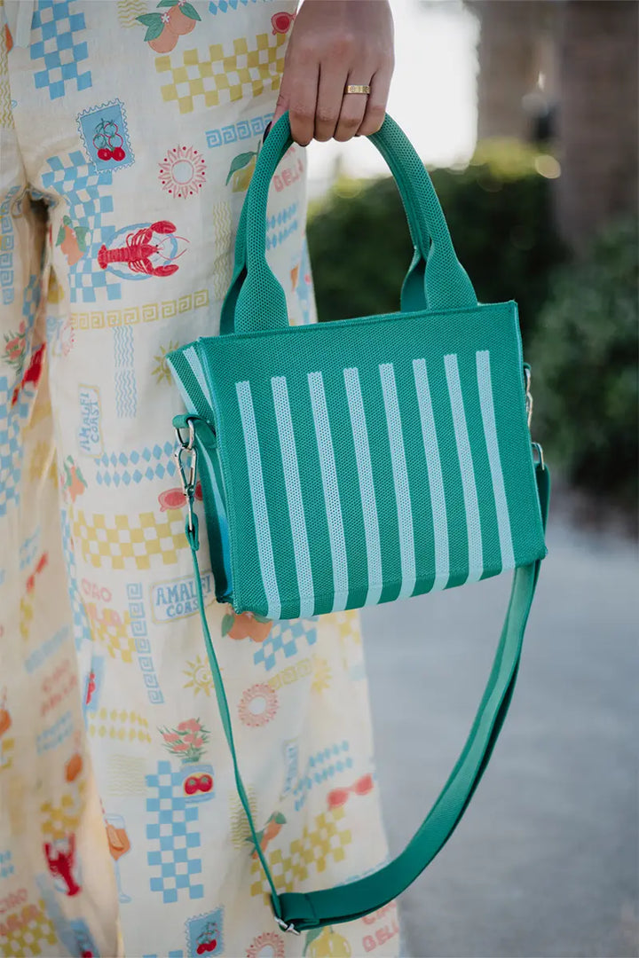 Woman in a colorful lobster and cherry print dress holding a green and mint stripe mesh mini tote bag with padded top handles and detachable crossbody strap, outdoors.