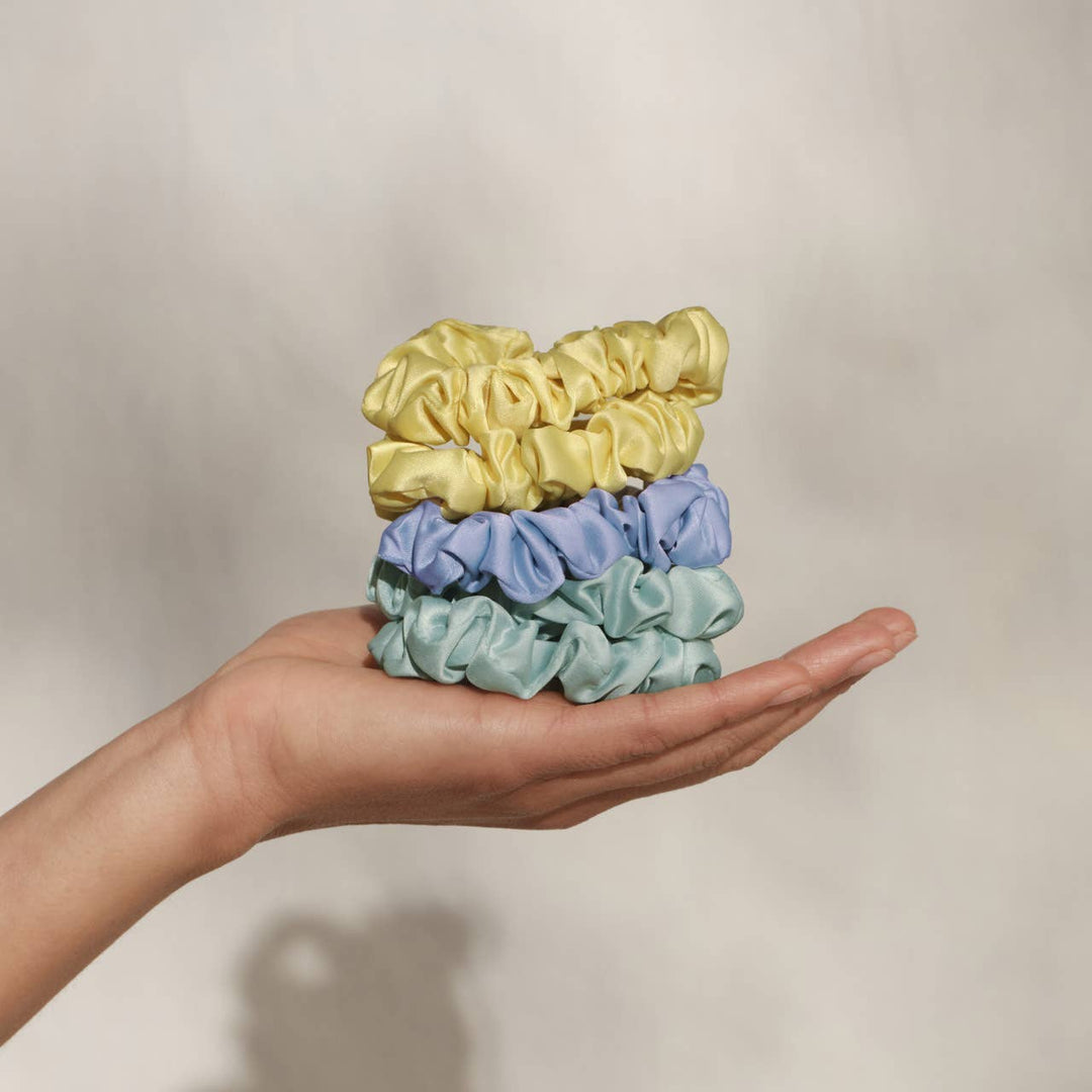 Hand holding a stack of three ruched satin scrunchies — butter yellow, periwinkle blue, and mint green — in an open palm against a neutral background.