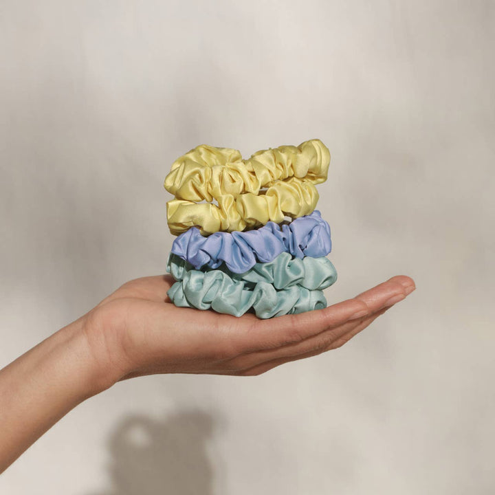 Hand holding a stack of three ruched satin scrunchies — butter yellow, periwinkle blue, and mint green — in an open palm against a neutral background.