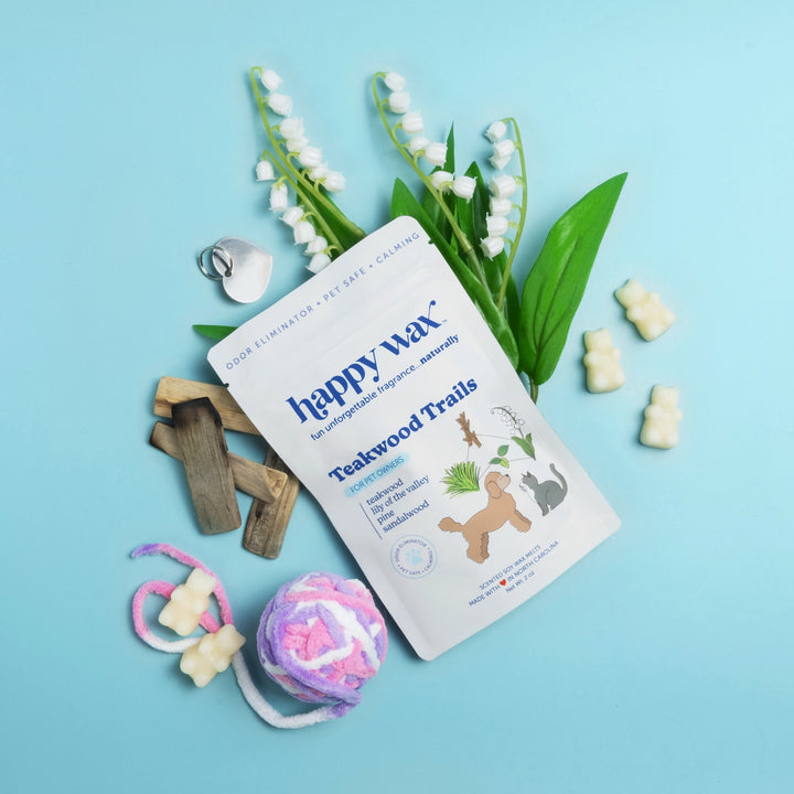 Happy Wax Teakwood Trails white pouch for pet owners with white bear-shaped wax melts, teakwood pieces, lily of the valley, and a pet toy on a light blue background.