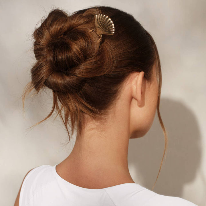 Back view of a woman with brown hair in a twisted bun secured with a gold scallop shell hair pin, wearing a white top.
