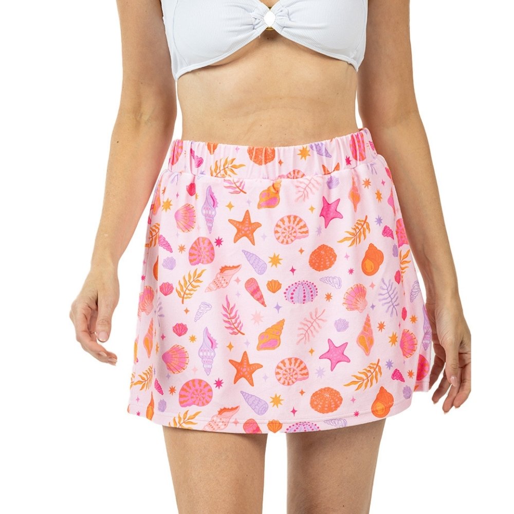 Front view of a pink seashell and starfish print elastic-waist mini skirt with pink, orange, and purple shell and botanical motifs, styled with a white knotted crop top.