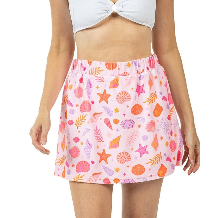 Front view of a pink seashell and starfish print elastic-waist mini skirt with pink, orange, and purple shell and botanical motifs, styled with a white knotted crop top.