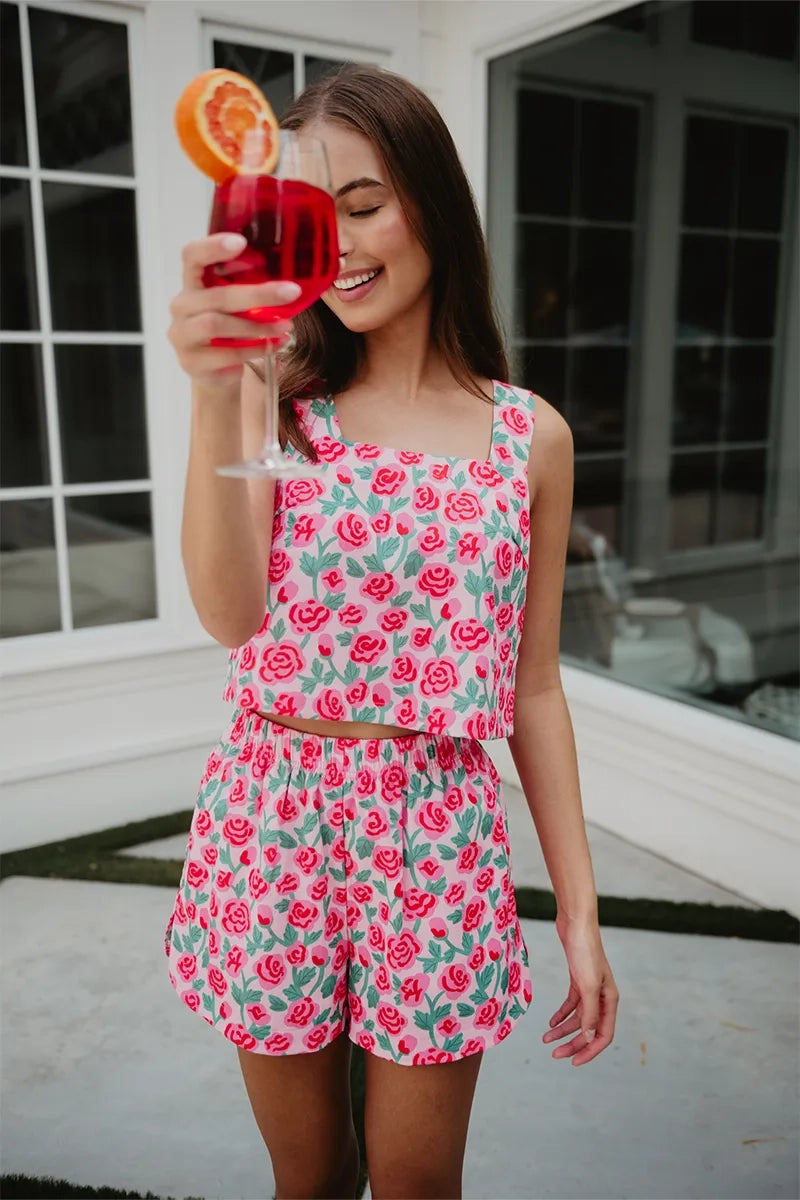 Smiling woman outdoors holding a red cocktail up to her face, wearing a pink rose print square-neck crop top and matching shorts set.
