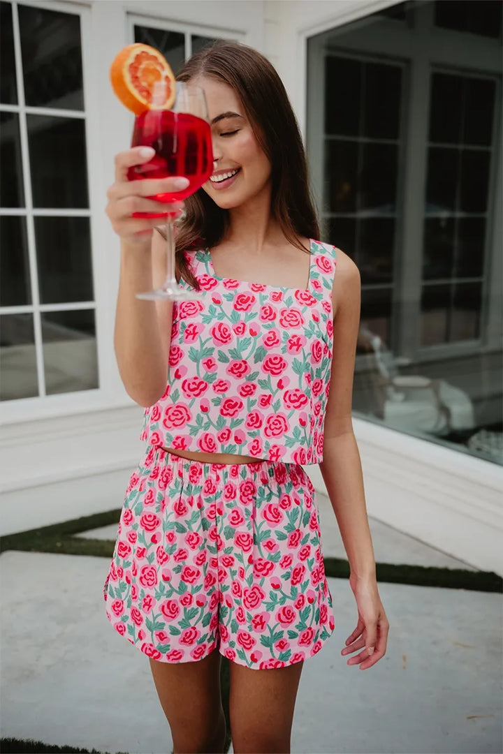 Smiling woman outdoors holding a red cocktail up to her face, wearing a pink rose print square-neck crop top and matching shorts set.