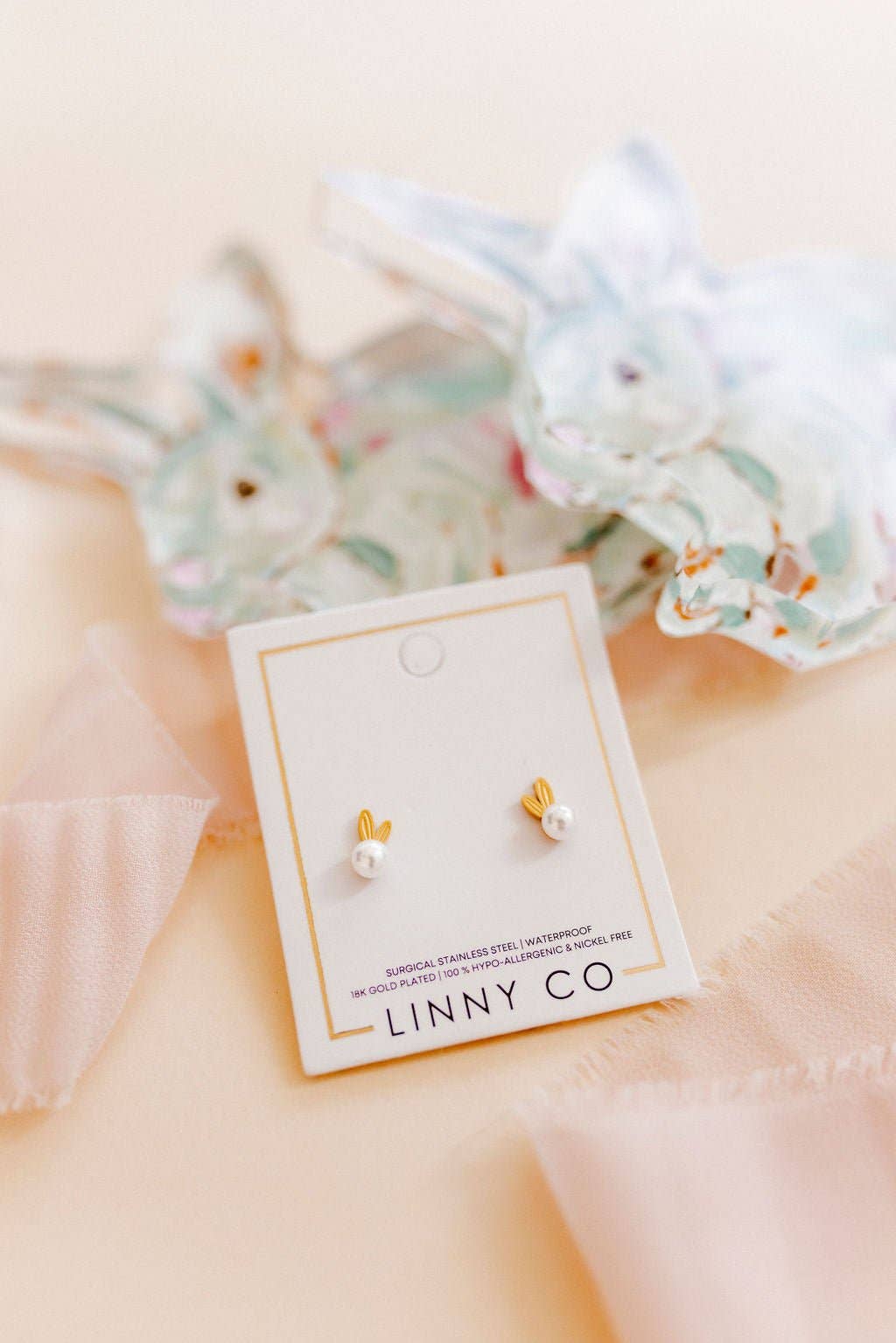 Linny Co gold bunny ear and white pearl stud earring card styled on a blush surface with soft blue floral and ribbon accents.