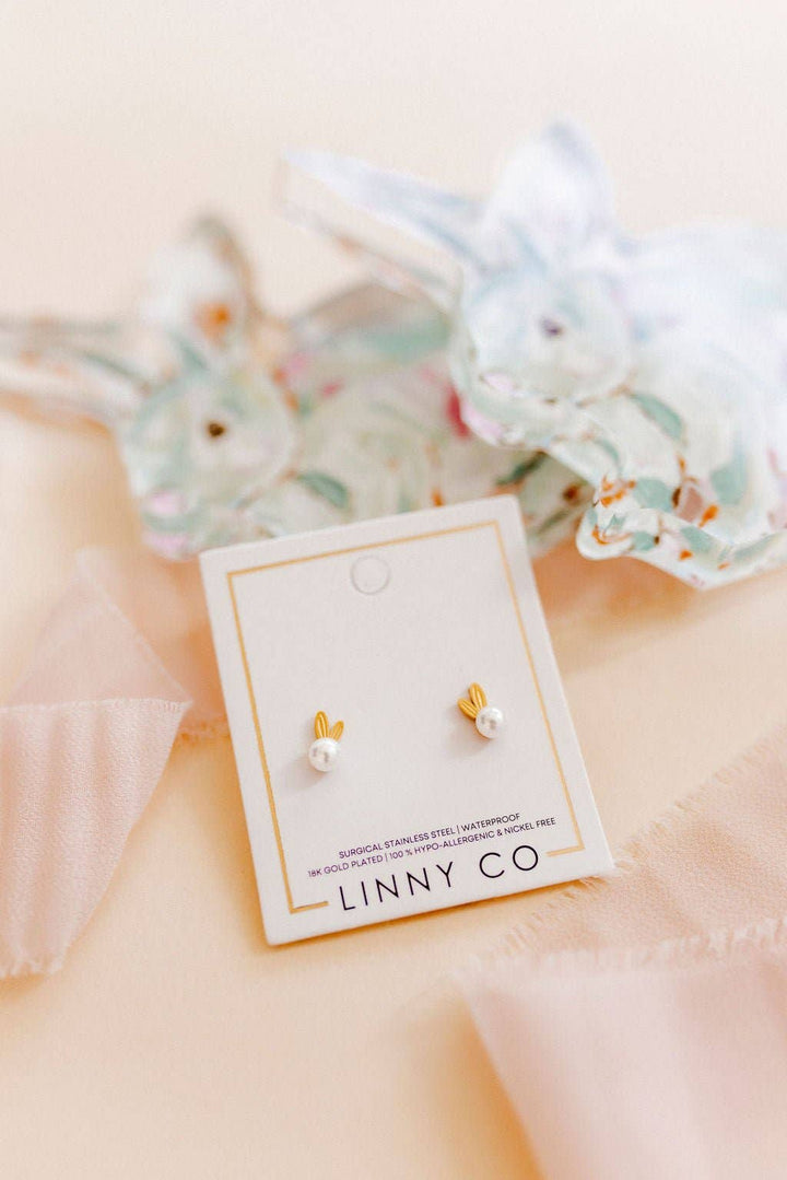 Linny Co gold bunny ear and white pearl stud earring card styled on a blush surface with soft blue floral and ribbon accents.