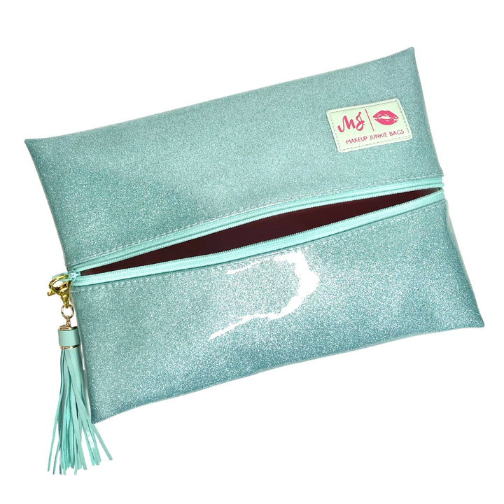 Makeup Junkie Bags mint glitter foldover zip makeup bag partially open showing burgundy interior lining with mint tassel zipper pull and branded patch, front view on white background