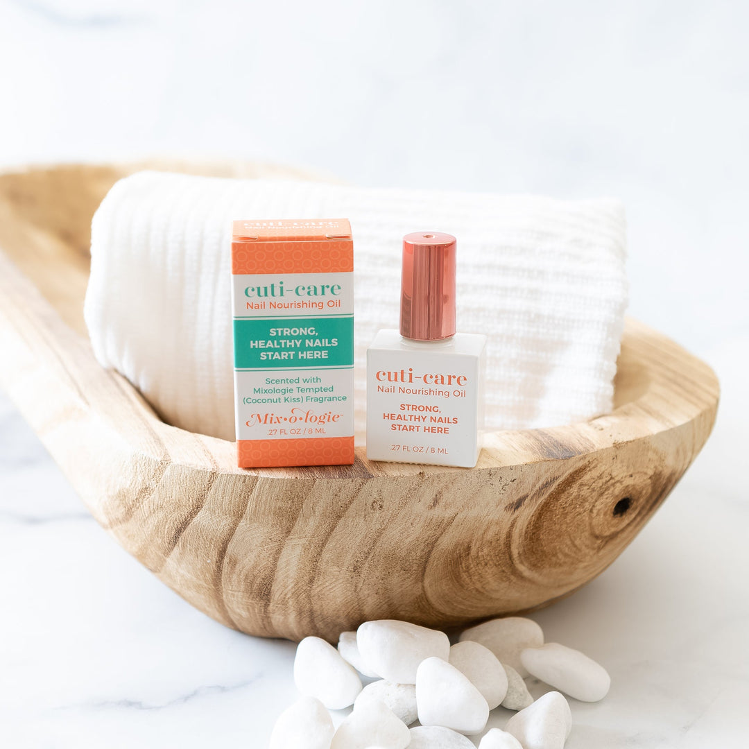Mixologie Cuti-Care Nail Nourishing Oil, white bottle with rose gold cap and orange box displayed in a rustic wood oval bowl with white river rocks and folded white towel