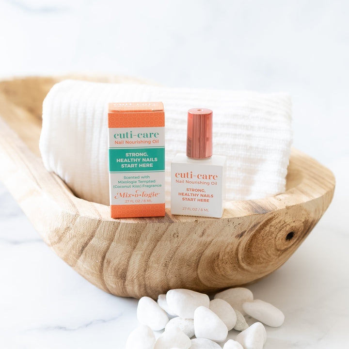 Mixologie Cuti-Care Nail Nourishing Oil, white bottle with rose gold cap and orange box displayed in a rustic wood oval bowl with white river rocks and folded white towel