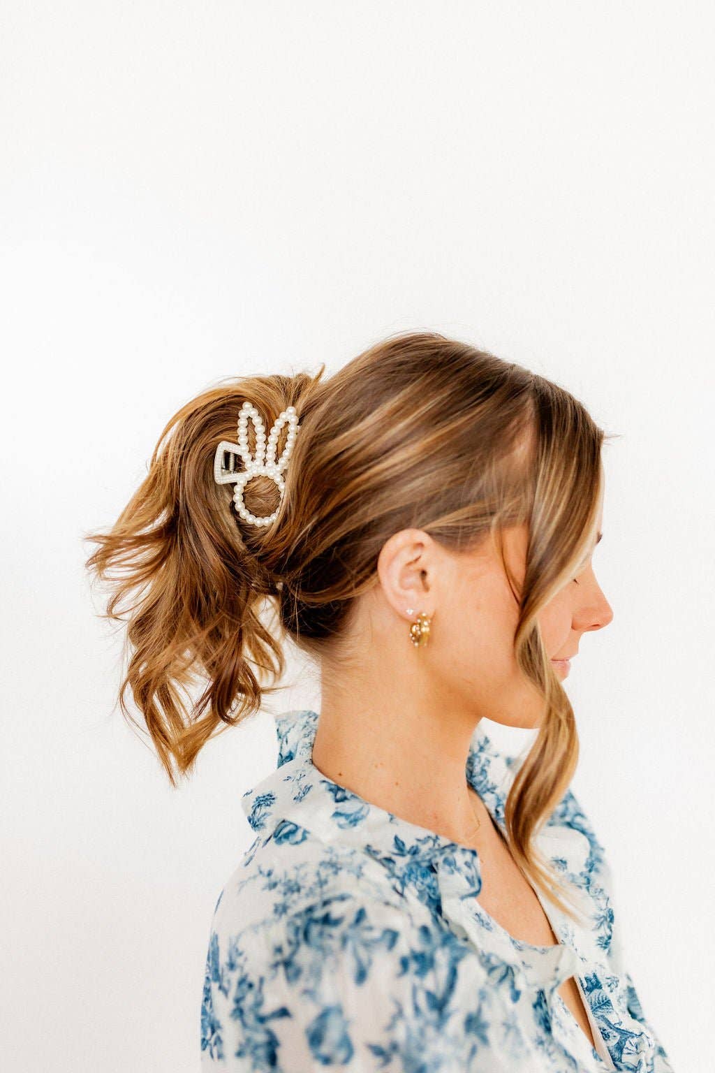 Side profile of woman wearing a pearl beaded bunny ear claw clip in a loose updo, styled with a blue floral top and gold flower stud earrings.