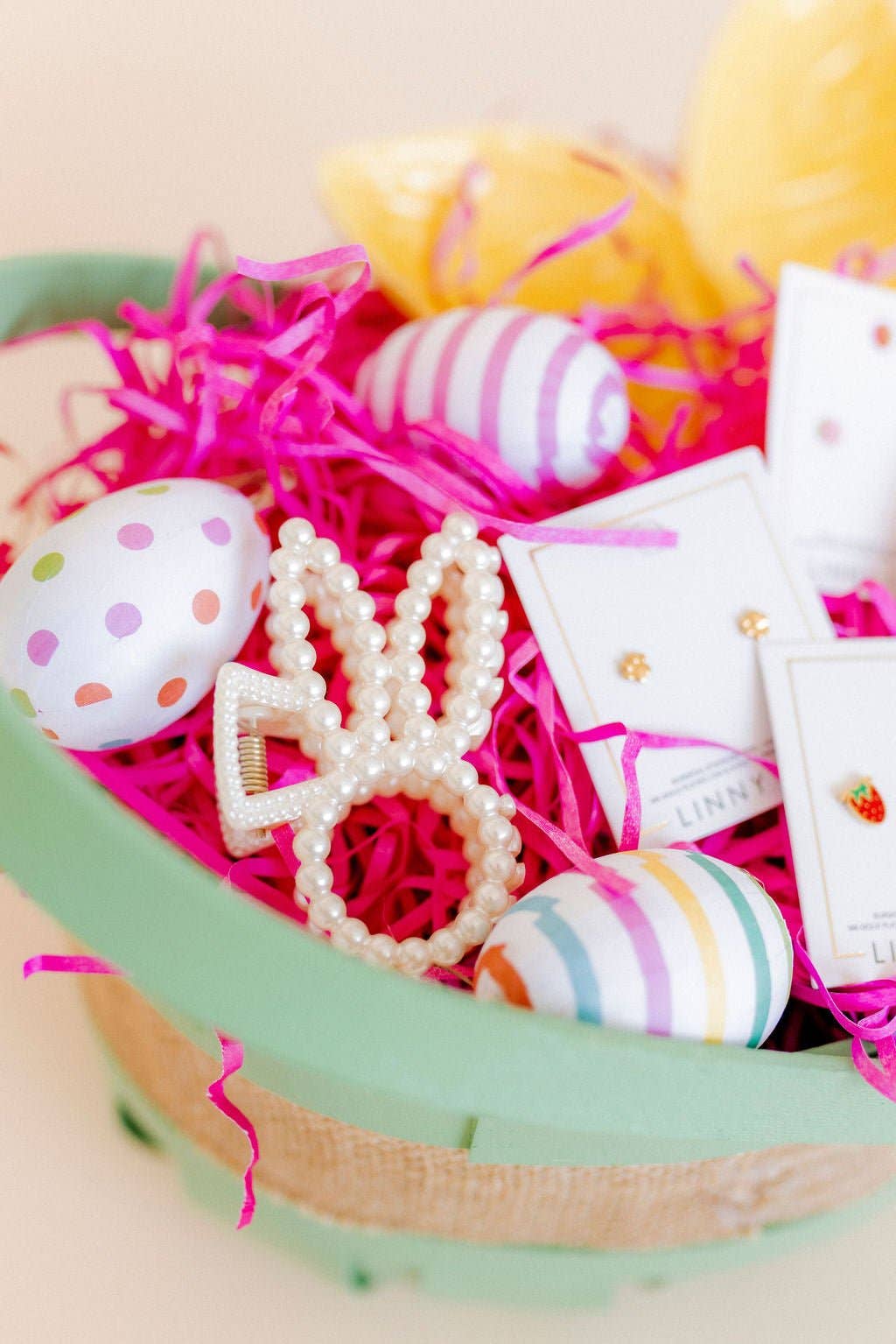Pearl beaded bunny ear claw clip nestled in a mint green Easter basket filled with hot pink shredded paper, polka dot and striped Easter eggs, and Linny Co earring cards.