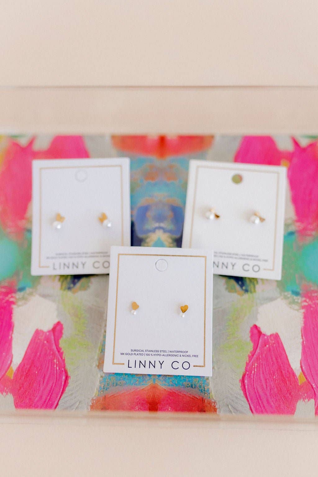 Three Linny Co gold bunny ear and pearl stud earring cards displayed on a colorful abstract painted surface.