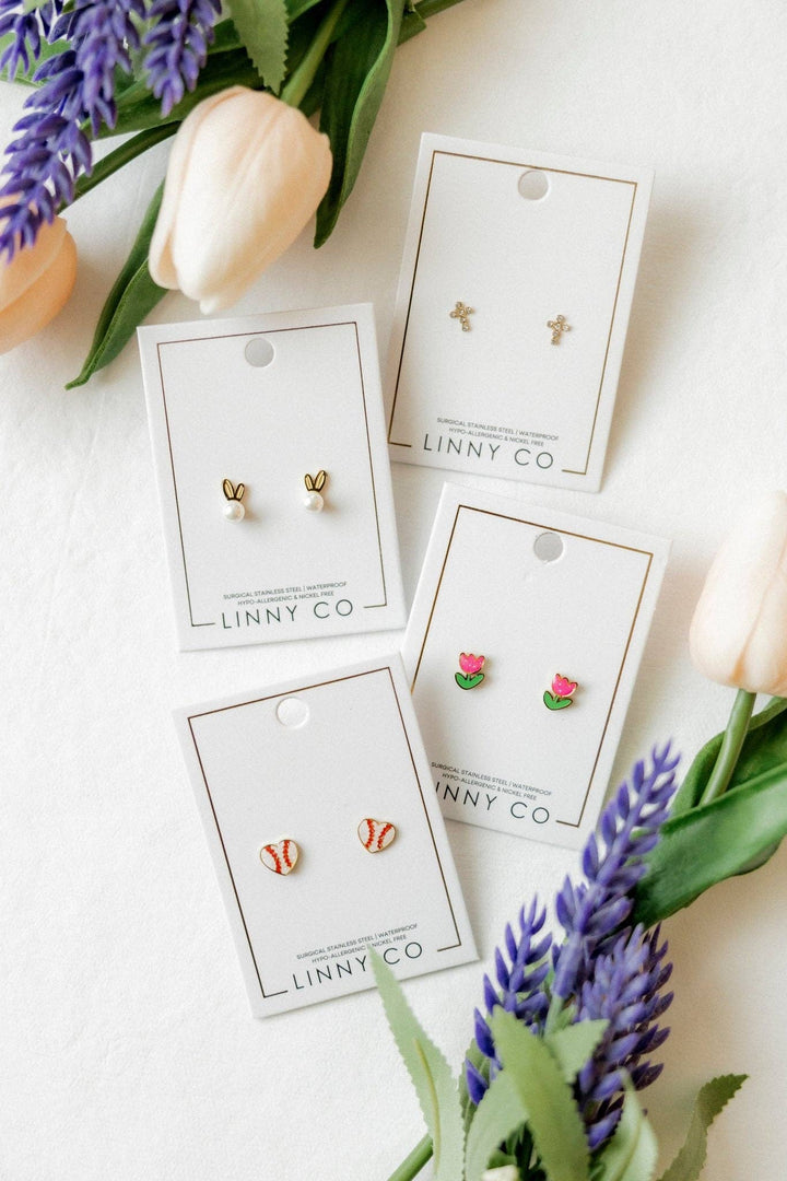 Four Linny Co stud earring cards flat lay surrounded by white tulips and purple lavender — gold bunny ear pearl studs, gold cross studs, pink tulip enamel studs, and red and white baseball enamel studs.