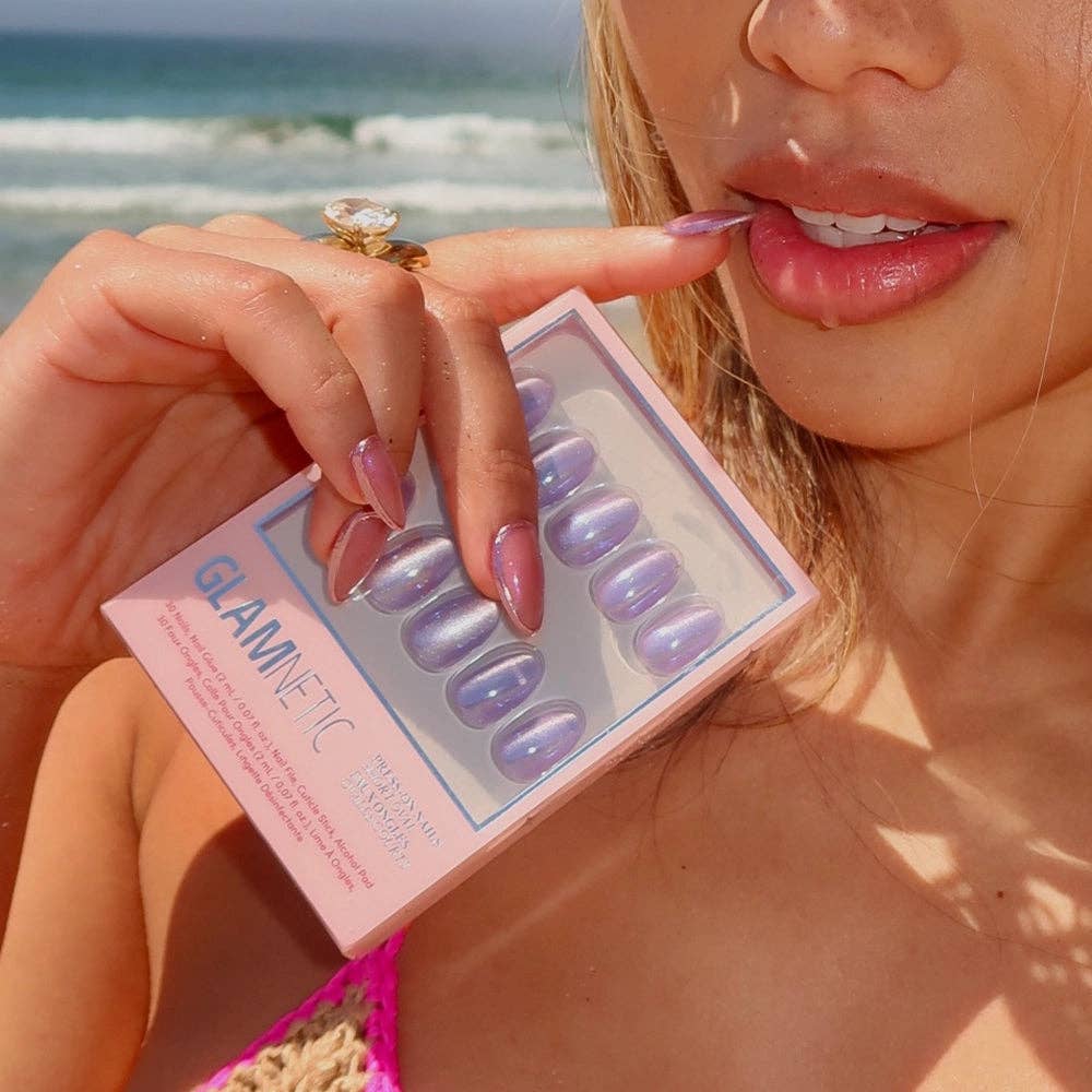 Woman in a pink bikini at the beach holding Glamnetic Mermaid's Tail Press-On Nails pink packaging, wearing iridescent lavender chrome short oval press-on nails and a gold cocktail ring