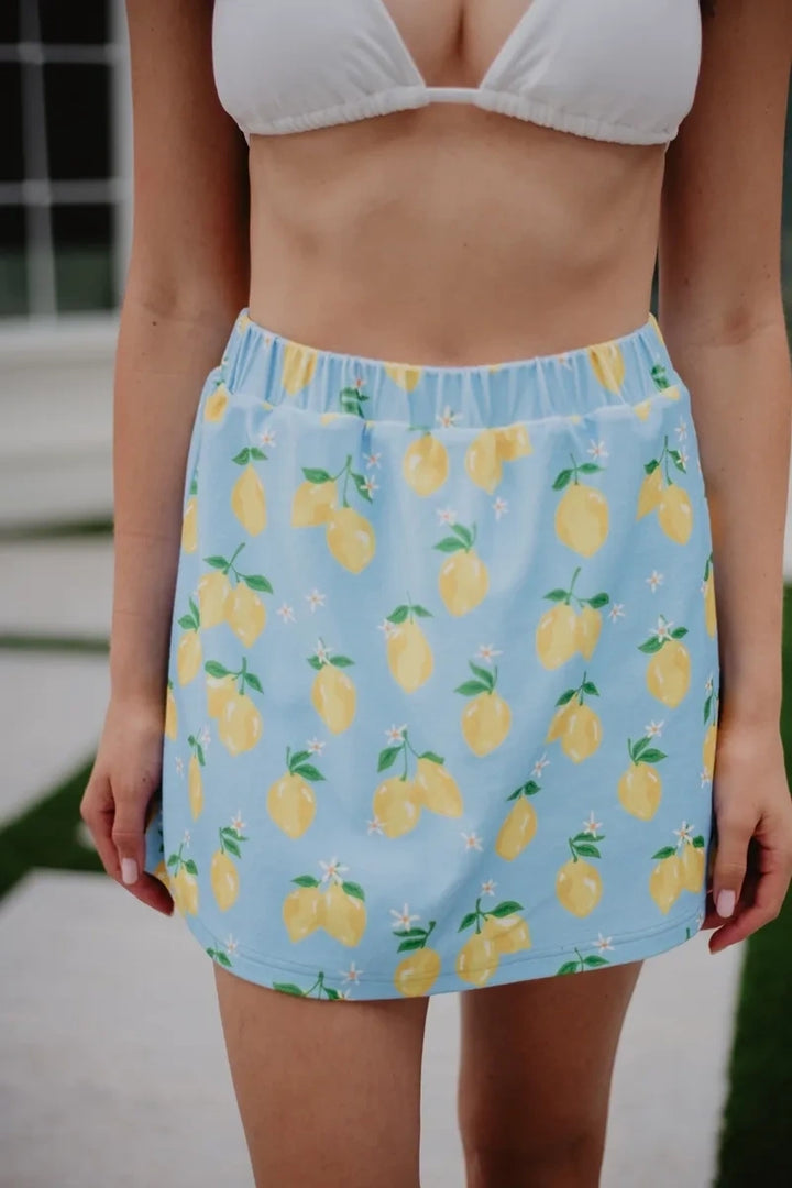 Front view of woman in a white bikini top and light blue lemon print elastic-waist mini skirt with yellow lemons, green leaves, and white daisy accents, standing outdoors.