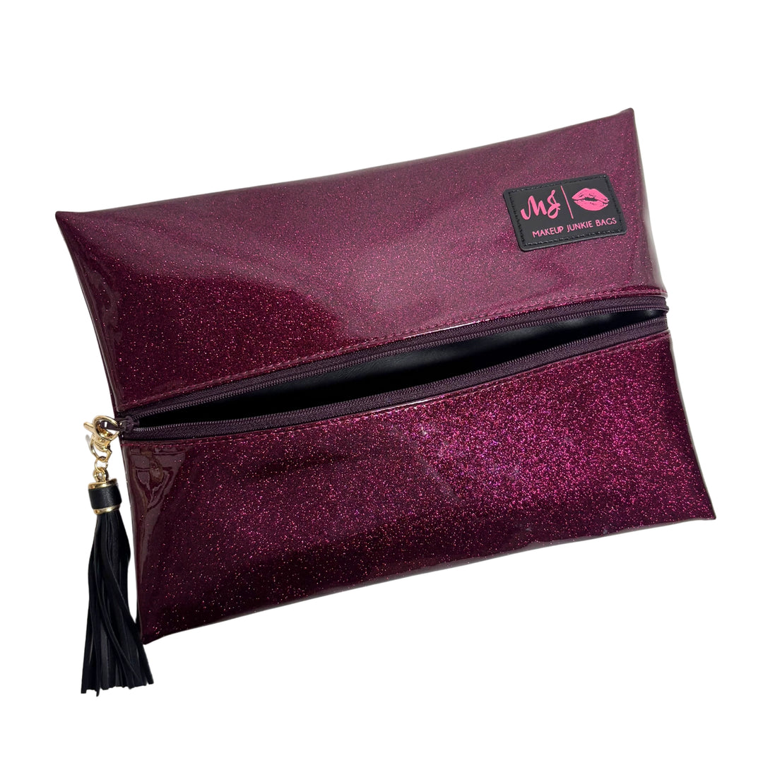 Makeup Junkie Bags magenta hot pink glitter foldover zip makeup bag partially open showing black interior lining with black tassel zipper pull and branded patch, front view on white background