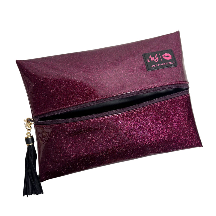 Makeup Junkie Bags magenta hot pink glitter foldover zip makeup bag partially open showing black interior lining with black tassel zipper pull and branded patch, front view on white background