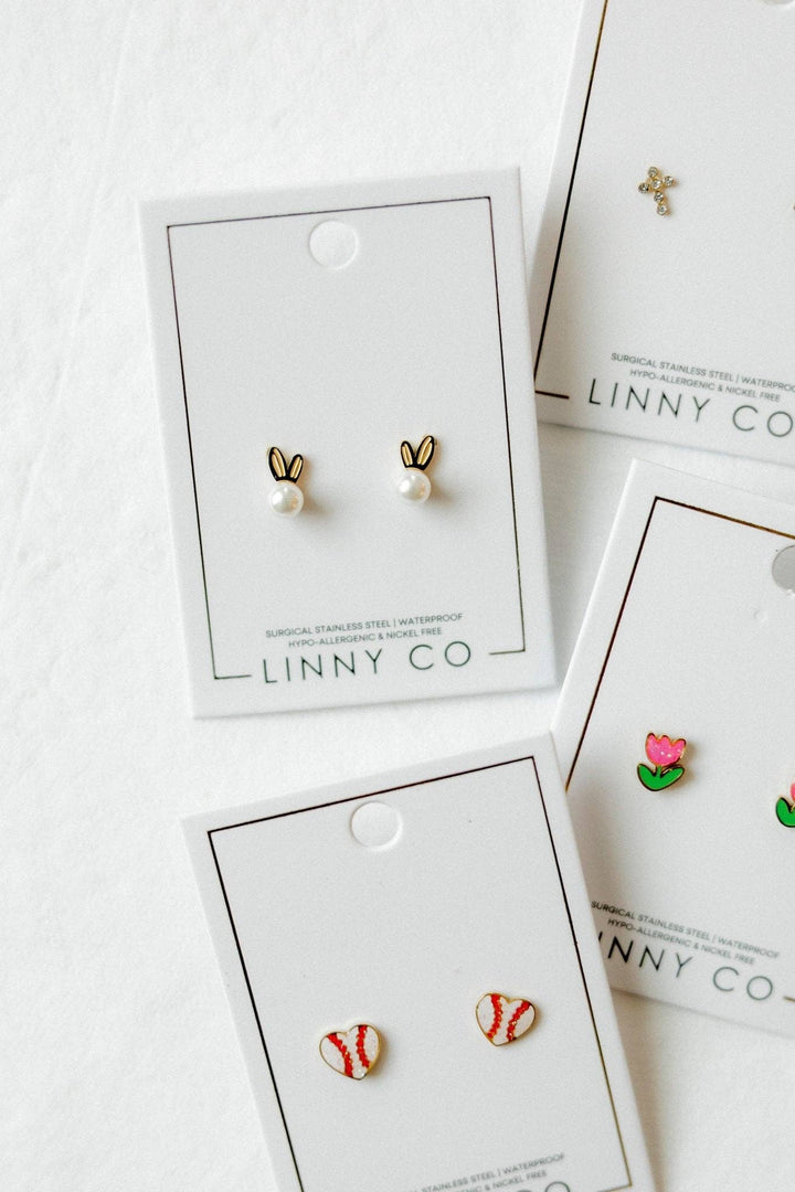 Linny Co stud earring cards flat lay on white background — gold bunny ear pearl studs and red and white baseball enamel studs in foreground, with gold cross and pink tulip enamel stud cards partially visible behind.
