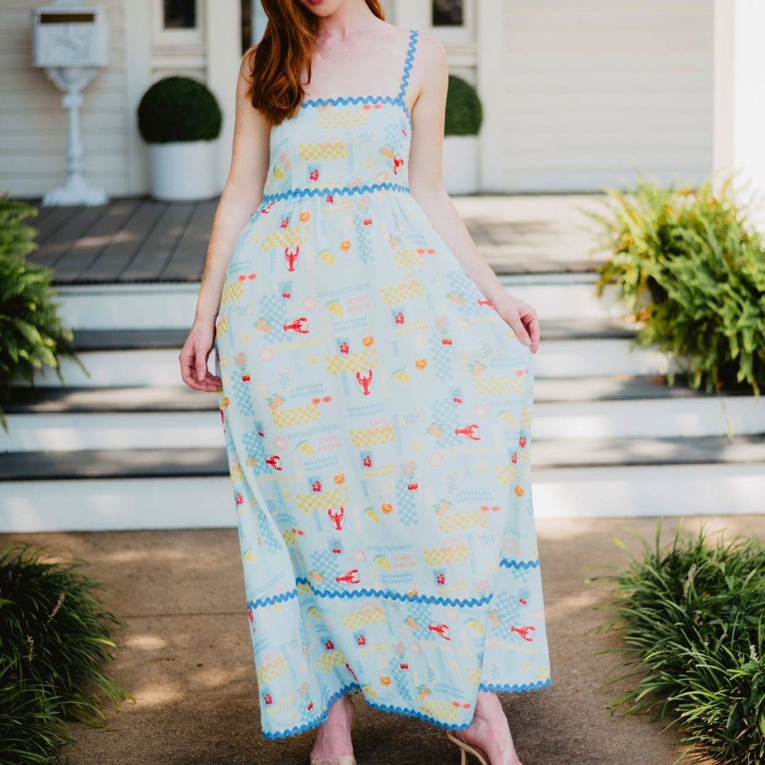 Front view of woman in a light blue lobster and summer print maxi dress with blue ric-rac trim at the neckline, waist, and hem, standing on a porch with greenery.