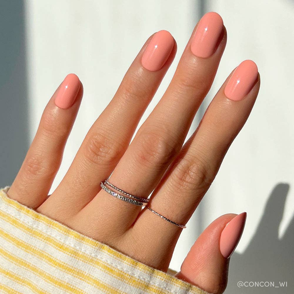 Close-up of hand wearing peach coral press-on nails with a delicate stacked ring, styled with a yellow striped sleeve