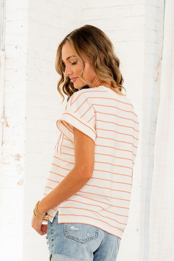 Back view of woman wearing a cream and orange stripe short sleeve top with cuffed sleeves and chest pocket.