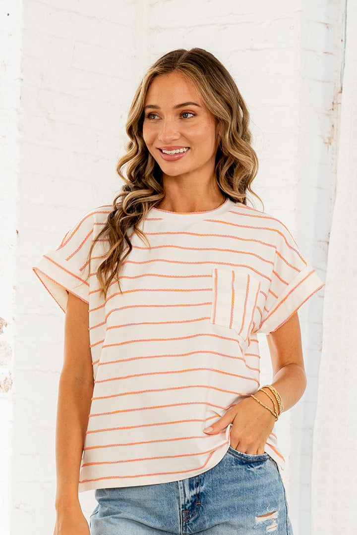 Woman wearing a cream and orange stripe short sleeve top with chest pocket and cuffed sleeves, paired with distressed jeans.
