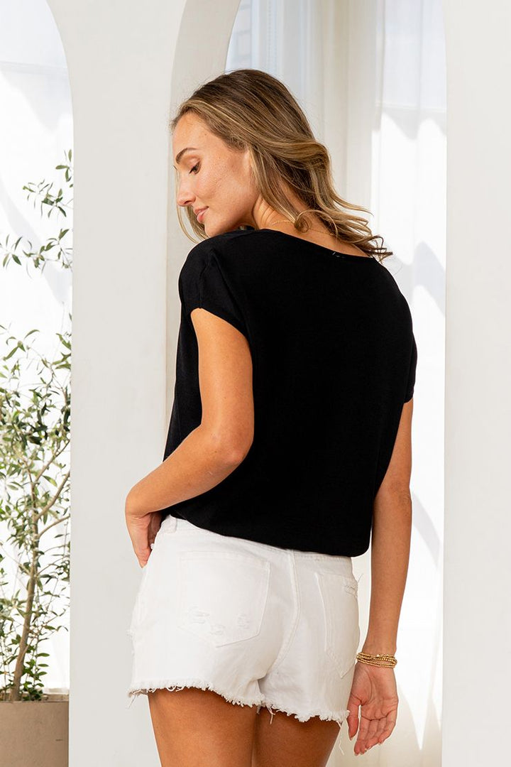 Back view of woman wearing a black v-neck short sleeve dolman top with distressed white shorts and gold bracelet.