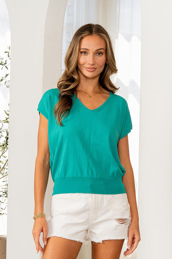 Woman in distressed white shorts and gold jewelry wearing a teal v-neck short sleeve dolman top, in front of a bright white window with soft natural light and greenery.