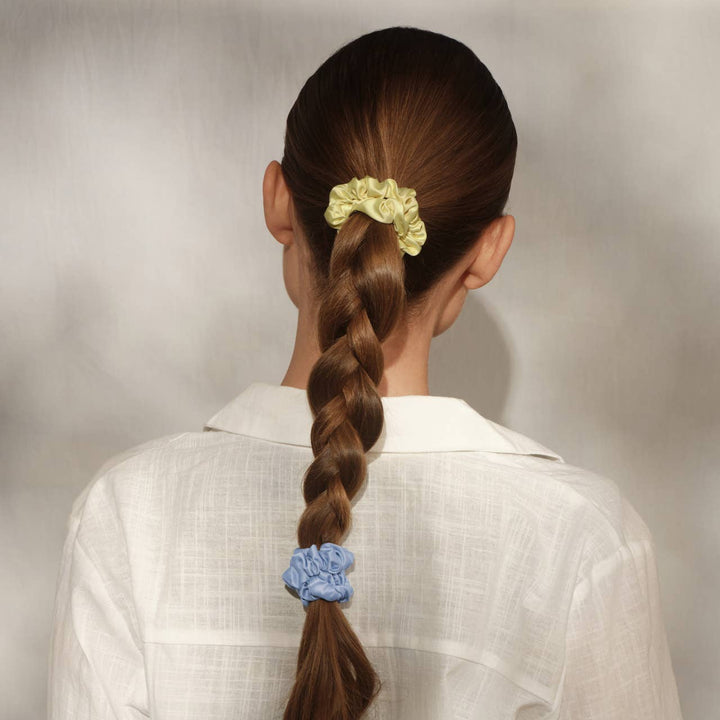 Back view of a woman with brown braided hair wearing a butter yellow satin scrunchie at the top of the braid and a periwinkle blue scrunchie at the end, in a white linen shirt.
