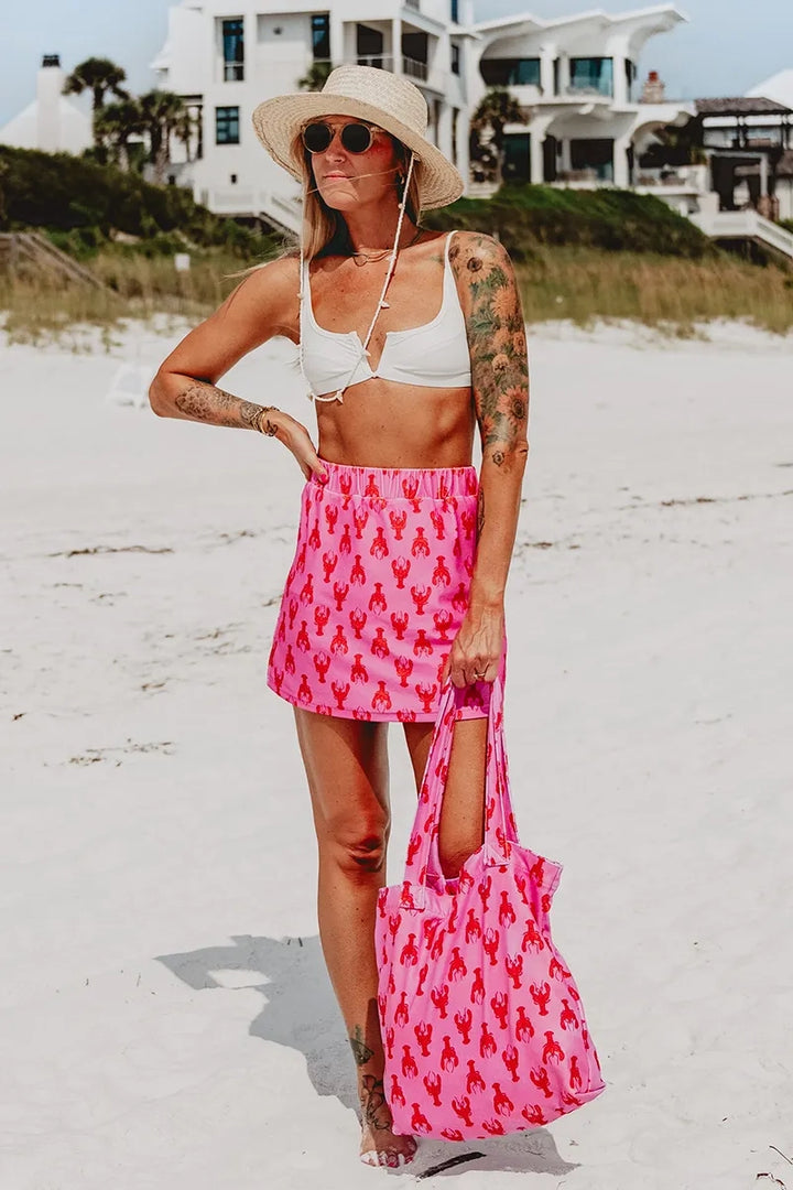 Woman in a straw hat, round sunglasses, and white bikini top wearing a pink lobster print elastic-waist mini skirt, carrying a matching pink lobster print tote bag, on a white sand beach.