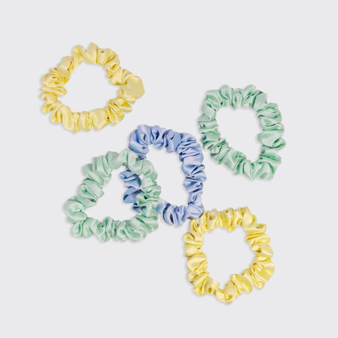 Flat lay of five ruched satin scrunchies in butter yellow, periwinkle blue, and mint green, spread out on a white background.