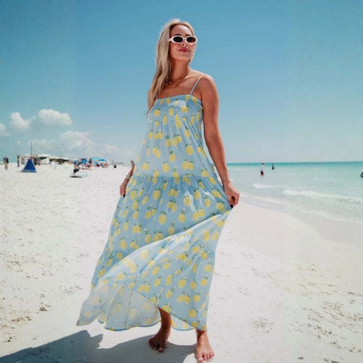 Woman in white oval sunglasses wearing a light blue lemon print smocked tiered maxi dress, holding the skirt as it flows in the breeze on a sunny beach.